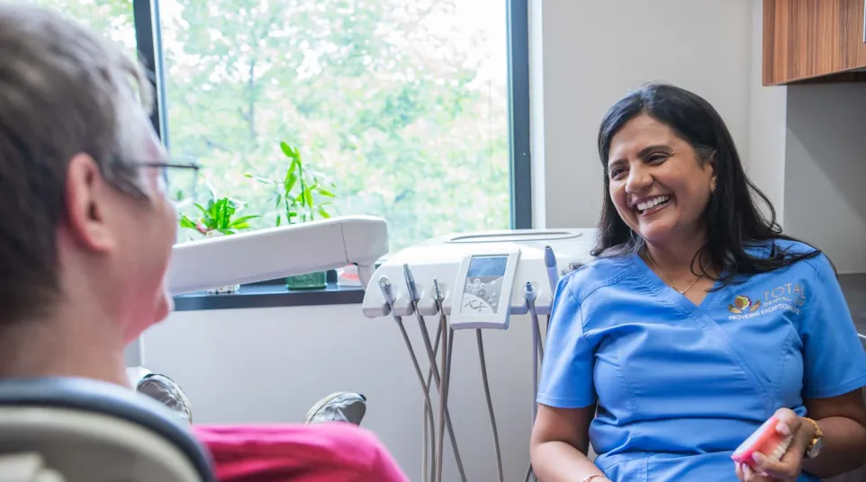 Dentist in blue scrubs smiling and talking to patient in dental office with equipment and window view.