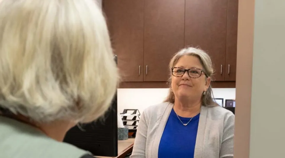 Woman in glasses and blue shirt consulting another woman about dental implants in an office setting.