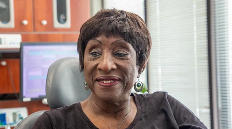 Smiling woman with short hair wearing a black shirt and cross necklace sitting in an office chair.