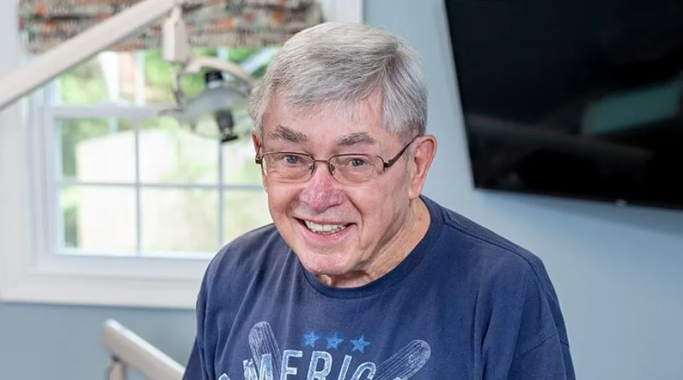 Elderly man wearing an American Baseball t-shirt smiling in a bright room with dental equipment.