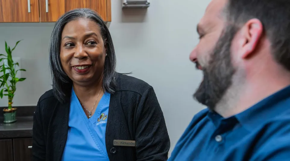 Female doctor in blue scrubs smiling and talking with a male patient in a medical office.