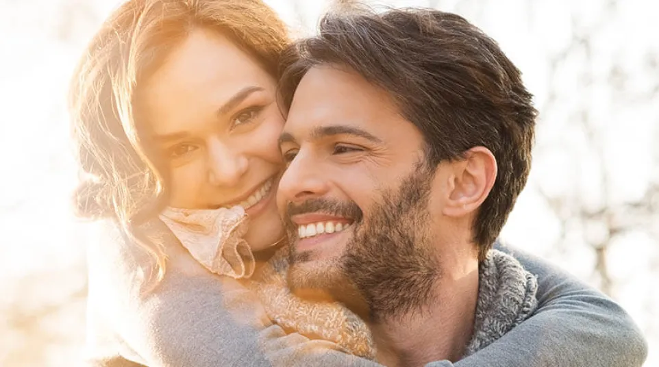 Happy couple embracing outdoors in cozy sweaters with warm sunlight in the background.
