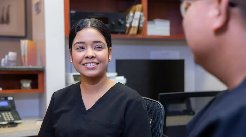 Smiling healthcare professional in black scrubs talking to a colleague in a medical office setting.