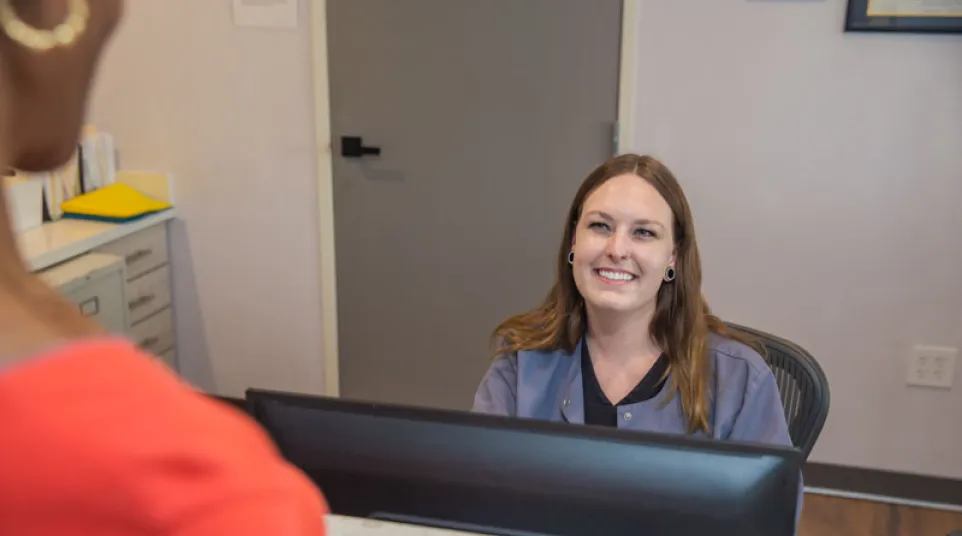 Smiling receptionist engaging with a visitor at a modern office front desk with computer and cabinets