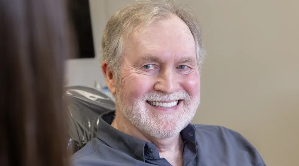 Smiling elderly man with gray beard sitting in a chair wearing a blue button-up shirt, facing a blurred person.