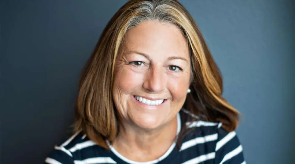 Smiling middle-aged woman with shoulder-length brown and blonde hair wearing a black and white striped shirt against dark background