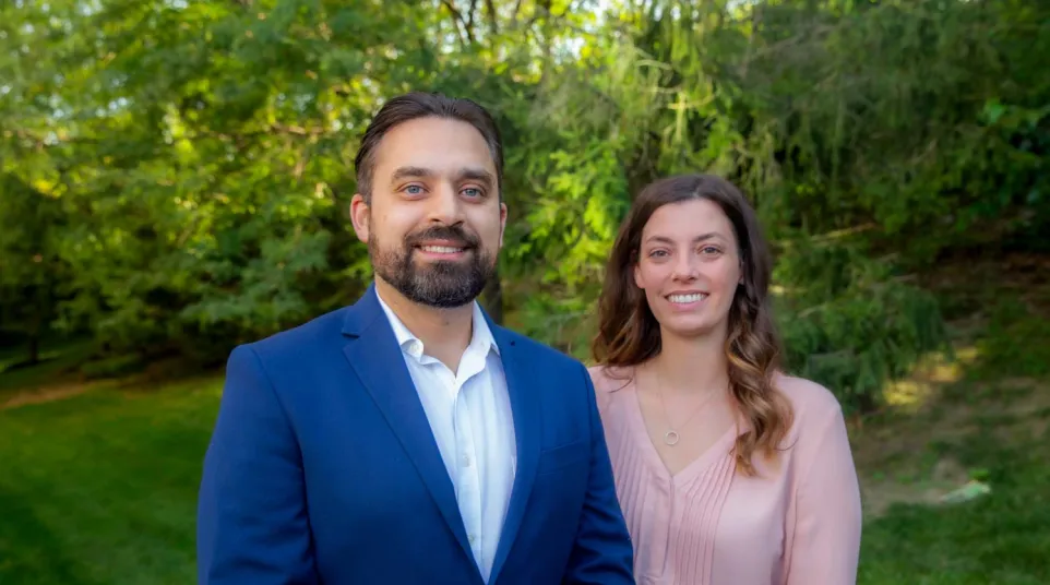 Smiling man in blue suit and woman in pink blouse standing outdoors with green trees and grass in background