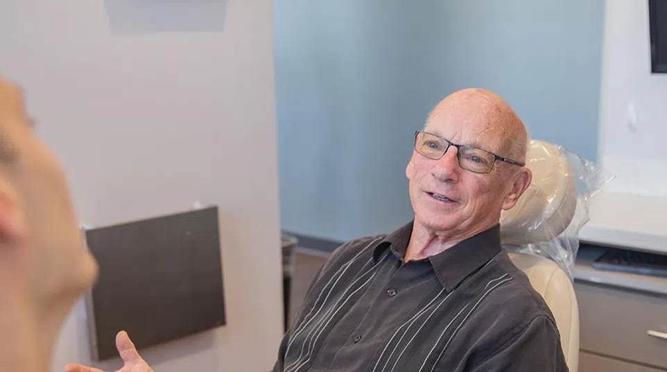 Elderly man with glasses sitting in a chair and talking to another person in a room with light walls.
