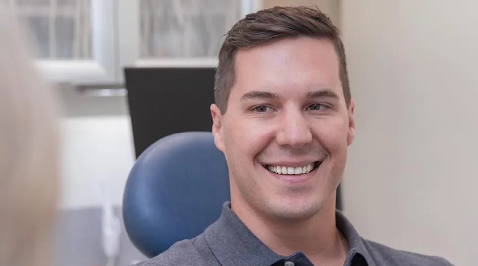 Young man in gray polo shirt smiling while sitting in a dental office chair during a consultation.