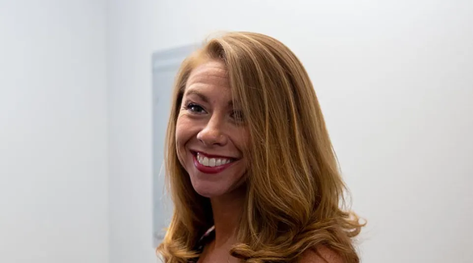 Smiling woman with long curly blonde hair wearing a patterned sleeveless dress against a plain light background