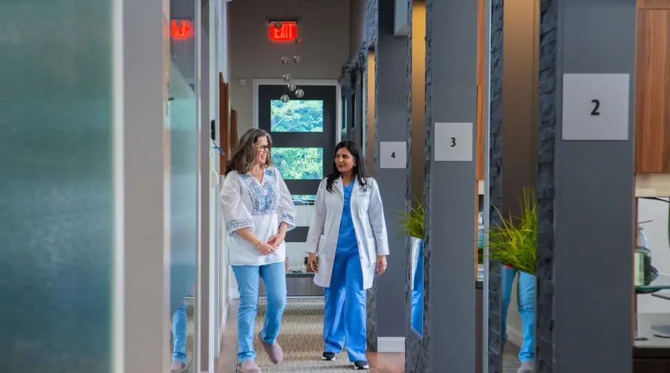 Female doctor in white coat walking and talking with female patient in a modern medical clinic hallway.