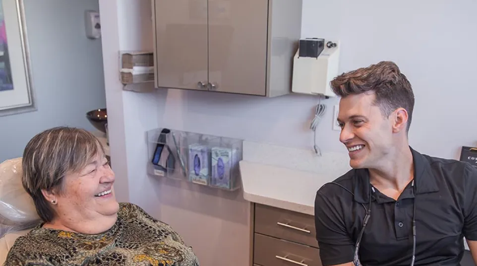 Smiling female dentist in black scrubs sitting at desk in a modern dental office with dental chair and equipment.