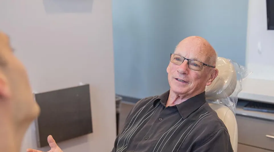 Elderly man in glasses talking and gesturing while sitting in a medical or dental office chair.