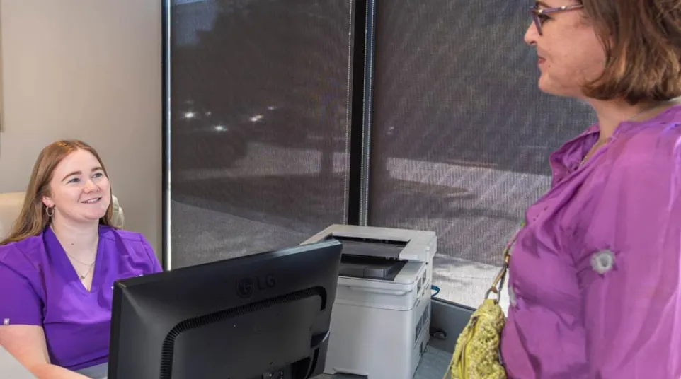 Smiling receptionist in purple scrubs assists woman in purple dress with yellow bag at front desk in office.