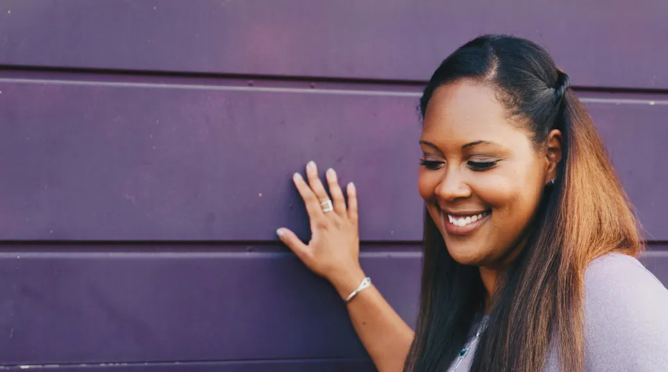 Smiling woman with long hair resting hand on purple wall, wearing casual light purple top and silver jewelry.