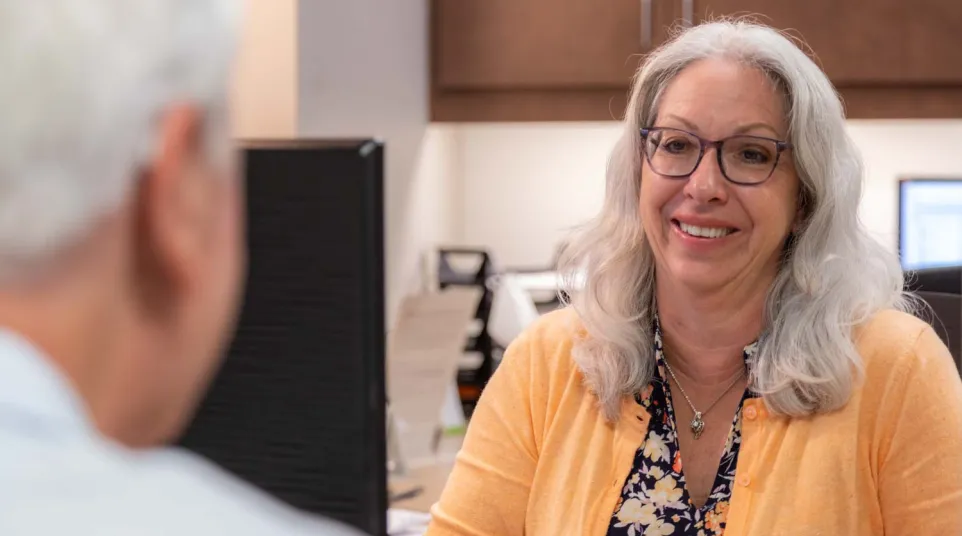 Smiling woman in glasses and orange cardigan talking to a man in an office setting.