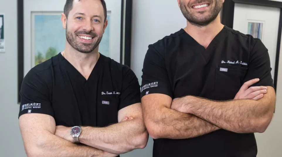 Two male doctors in black scrubs posing with arms crossed, smiling in a medical office setting.