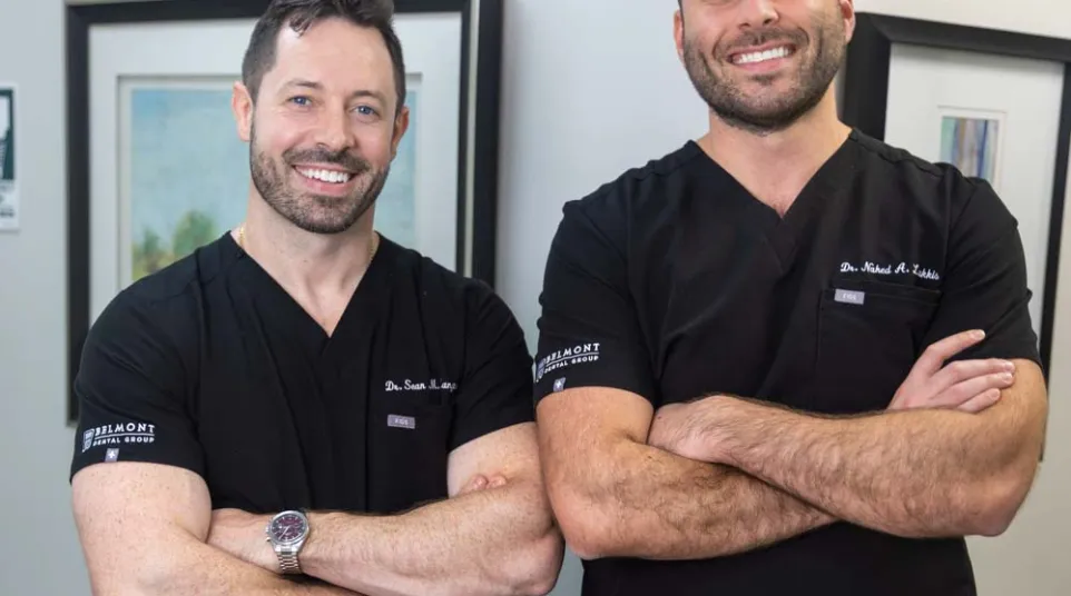 Two male doctors in black scrubs posing with arms crossed, smiling in a medical office setting.