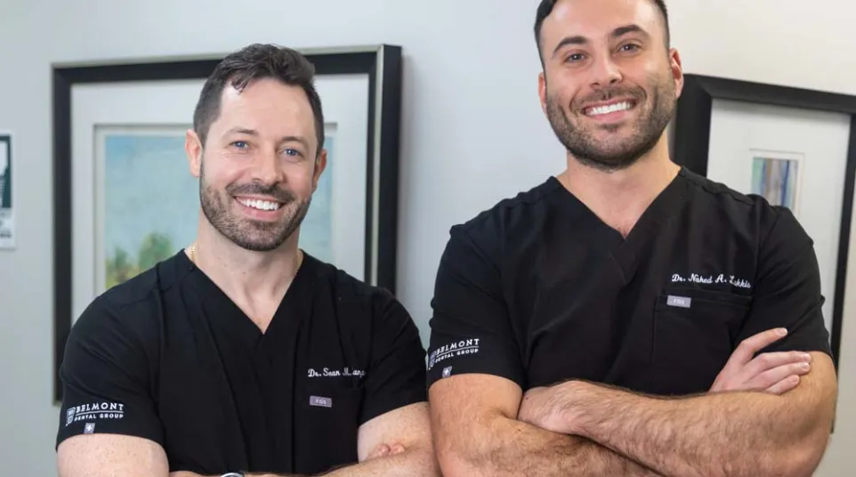 Two male doctors in black scrubs posing with arms crossed, smiling in a medical office setting.