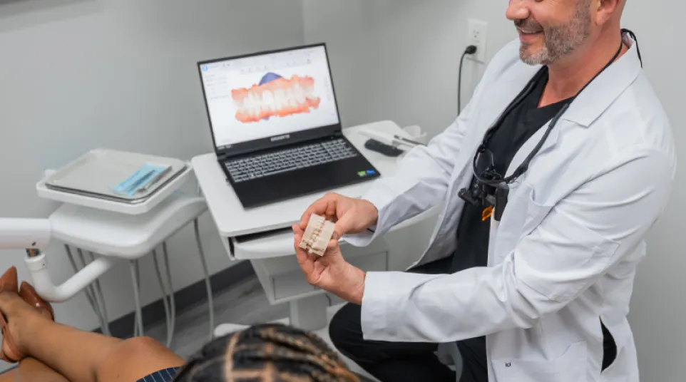 Dentist in white coat showing dental mold to patient in modern dental office with laptop and screen.