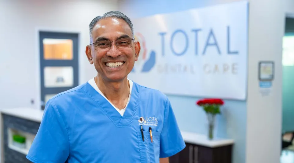 Smiling male dentist in blue scrubs standing in Total Dental Care clinic with a bright, welcoming atmosphere.