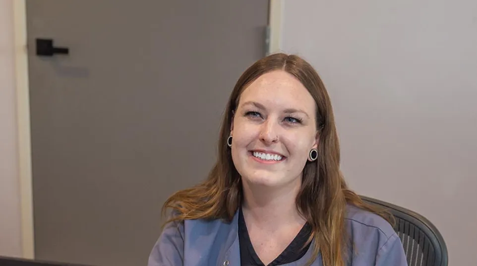 Smiling woman with brown hair in casual uniform sitting behind a desk with a computer monitor.
