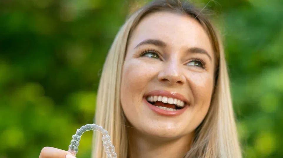 Smiling young woman holding a clear dental aligner outdoors with green blurred background