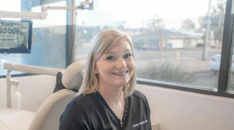 Smiling female dental professional in black scrubs sitting at a desk with dental chair and large windows behind her.