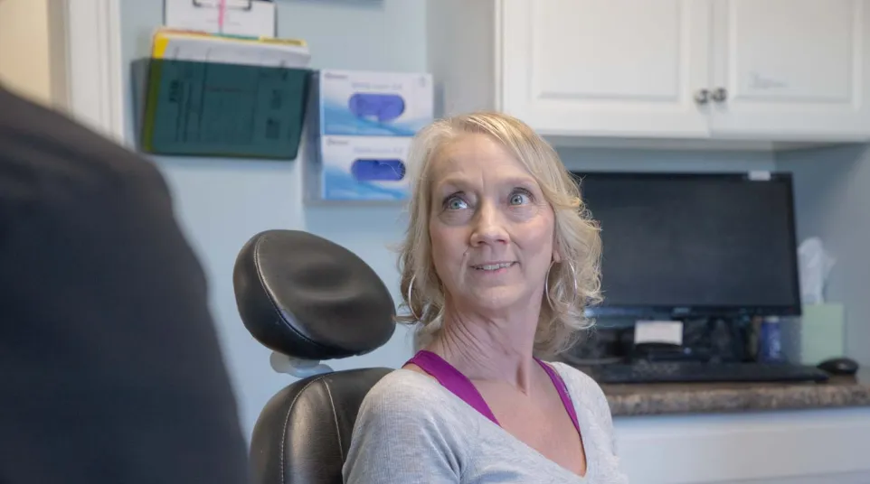 Middle-aged woman sitting in a dental chair looking toward dentist in a modern clinic room.