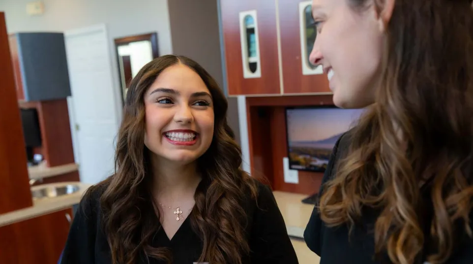 Two female healthcare professionals smiling and talking in a medical office with computer monitors and cabinets.