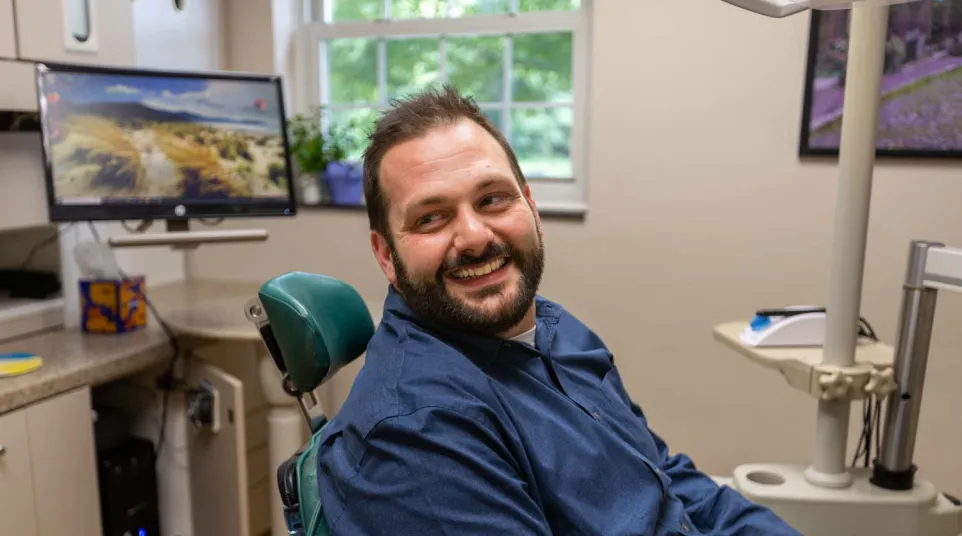 Smiling man sitting in a dental chair in a bright dental office with equipment and monitor visible.