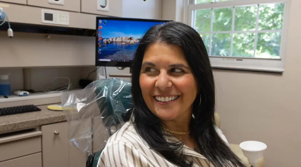 Smiling woman sitting in a dental chair in a bright dental office with a computer screen and window in background