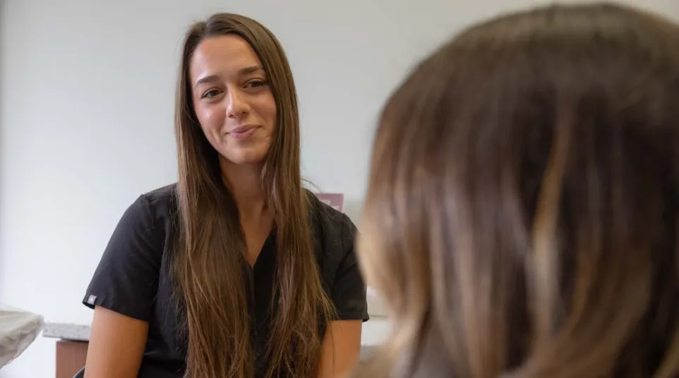 Smiling woman with long brown hair in black shirt conversing warmly with another person in indoor setting