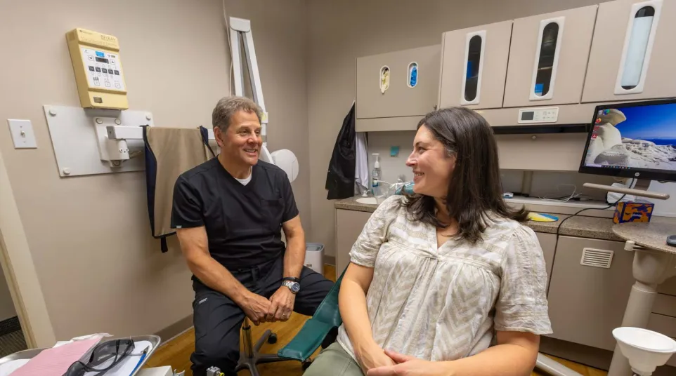 Dentist in black scrubs talking and smiling with a female patient in dental chair inside a clinic room.