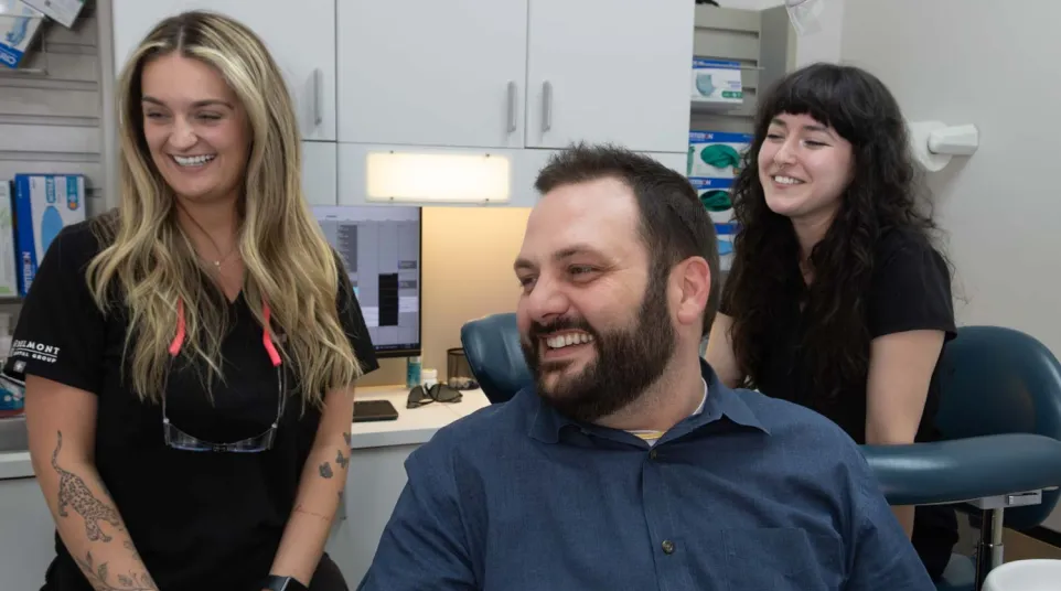 Smiling male patient and two female dental staff in a modern dental office with computer and medical supplies.