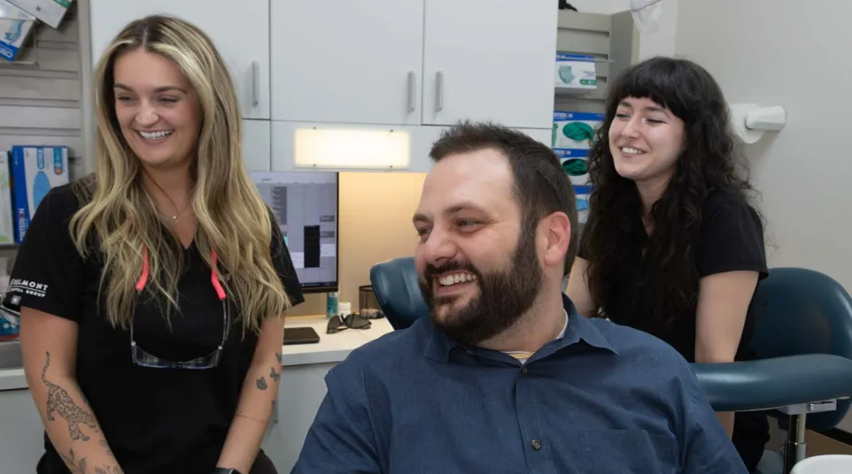 Smiling male patient and two female dental staff in a modern dental office with computer and medical supplies.
