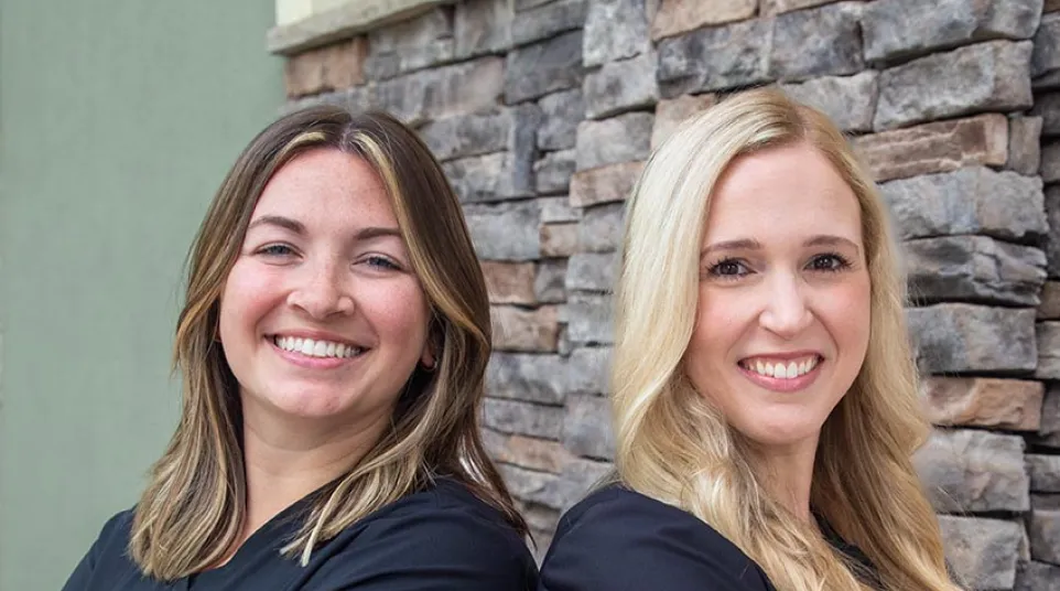 Two smiling women in black scrubs stand back-to-back with arms crossed in front of a stone wall.