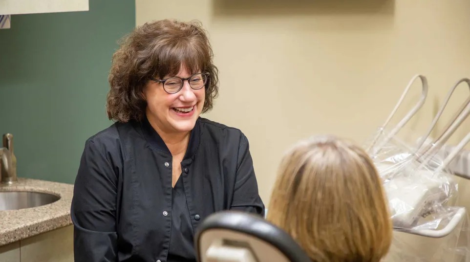 Smiling female dentist in black coat talking to female patient in dental office with equipment.