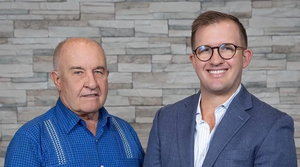 Older man in blue shirt and younger man in suit stand smiling against stone wall background.