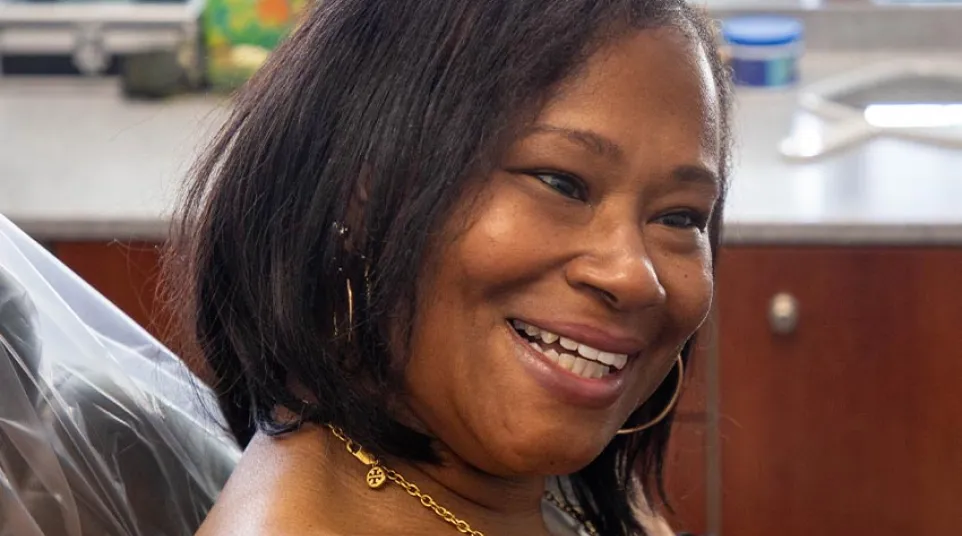 Smiling woman with shoulder-length hair wearing gold jewelry and a black top in a bright room.