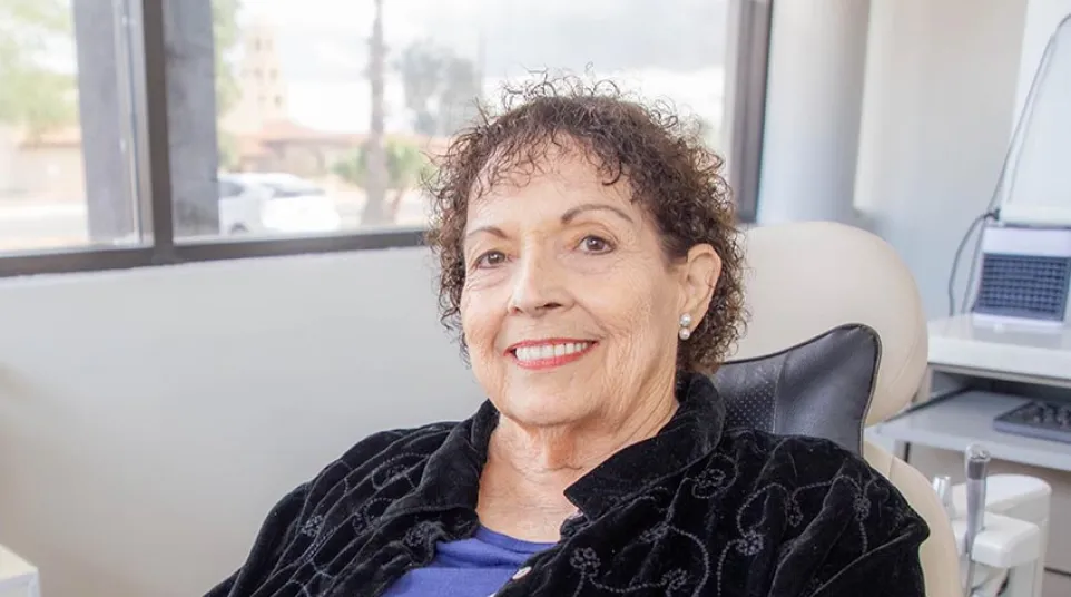 Smiling elderly woman sitting in a chair indoors wearing a black jacket and blue patterned shirt
