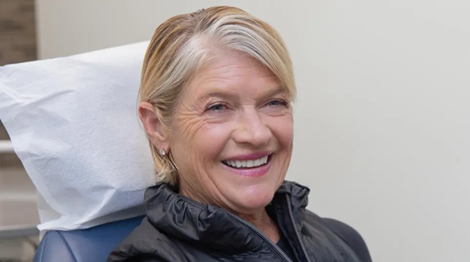 Smiling senior woman with short blonde hair sitting in a chair wearing a black vest indoors.