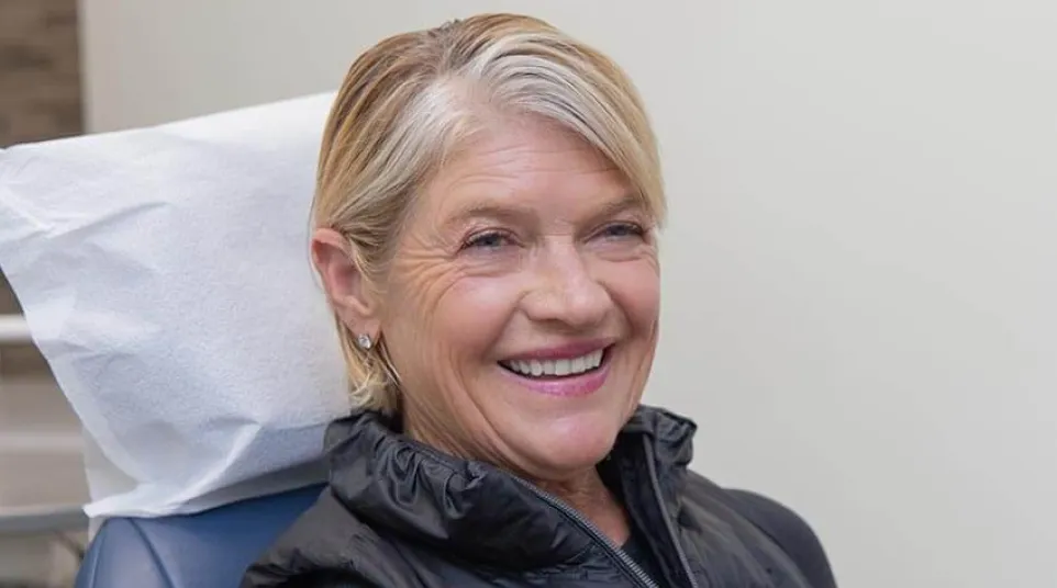 Smiling senior woman with short blonde hair sitting in a chair wearing a black vest indoors.
