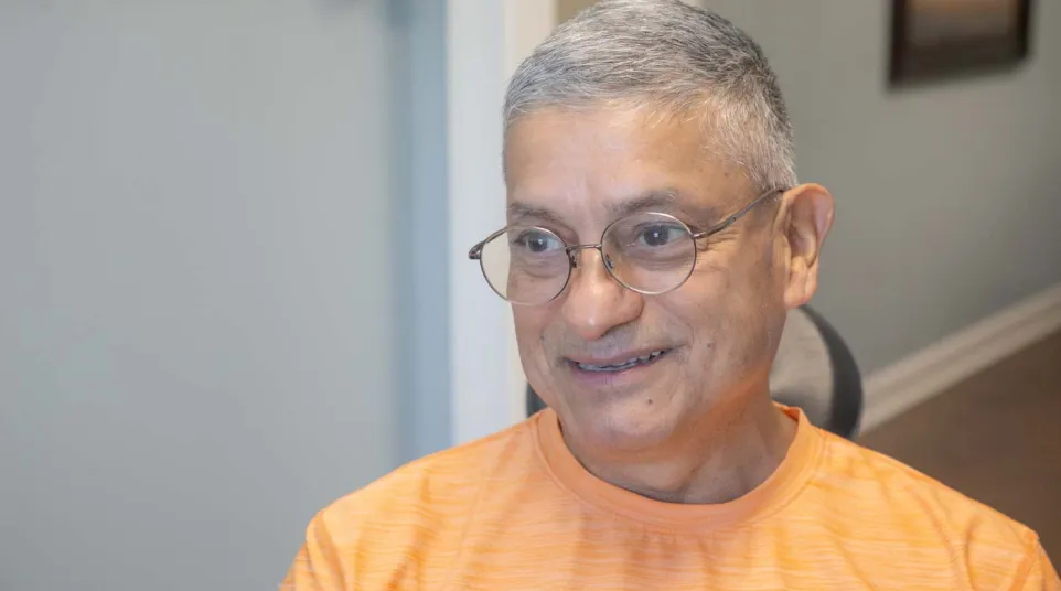 Smiling older man with gray hair and glasses wearing an orange shirt indoors in a home setting