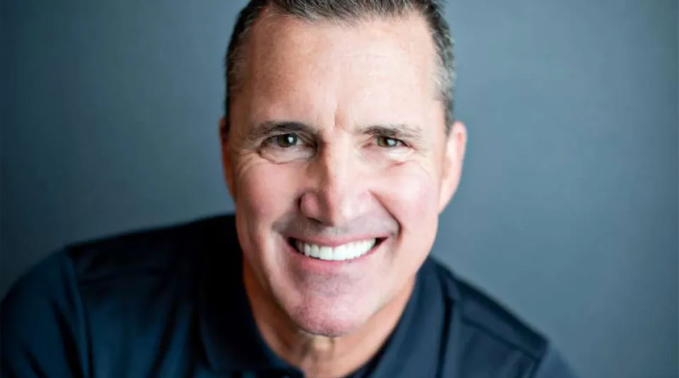 Smiling middle-aged man wearing a black polo shirt with bright white teeth against a gray background.