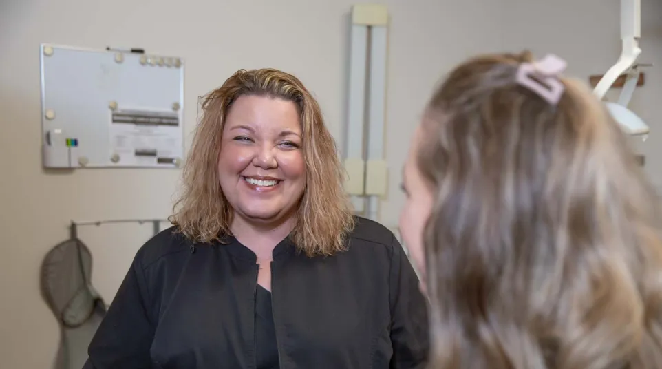 Smiling woman in black jacket talking to another person in indoor setting with medical or office equipment.