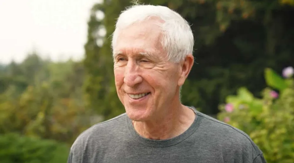 Elderly man with white hair smiling outdoors with blurred green foliage background