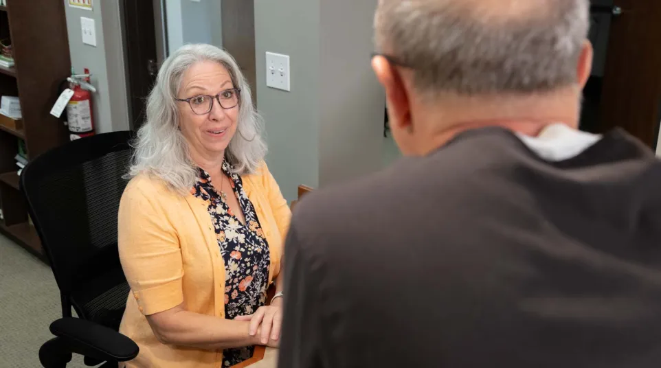 Mature woman in yellow cardigan talking to man at office desk, engaged in conversation during meeting