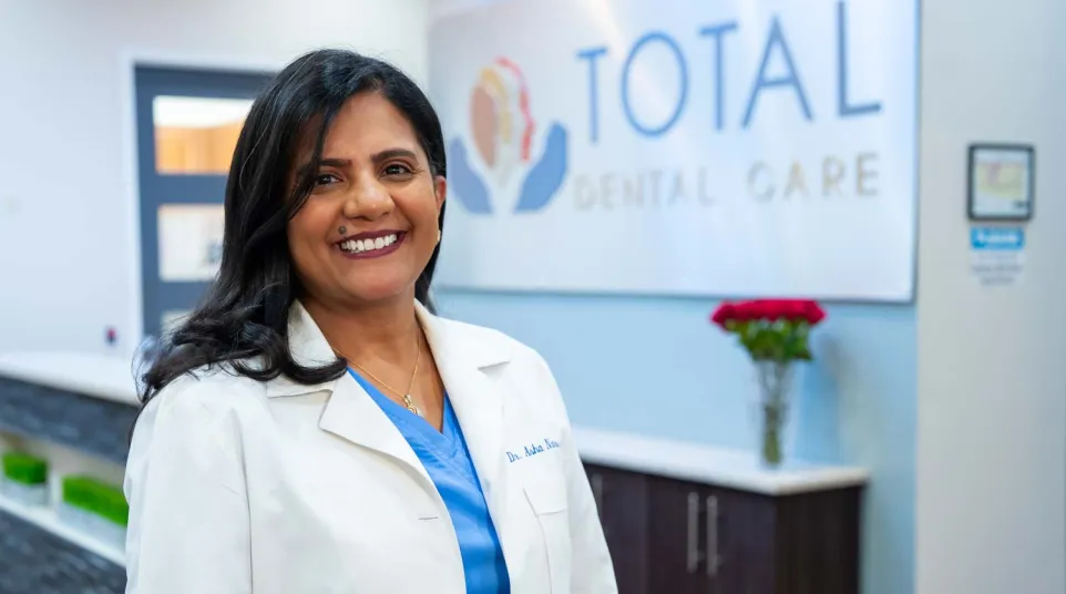 Smiling female dentist in white coat standing inside dental clinic lobby with Total Dental Care sign behind her.