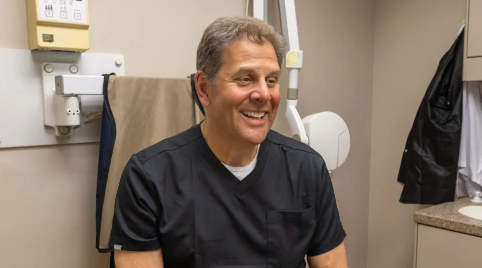 Smiling male medical professional in black scrubs sitting in a medical office with equipment in the background